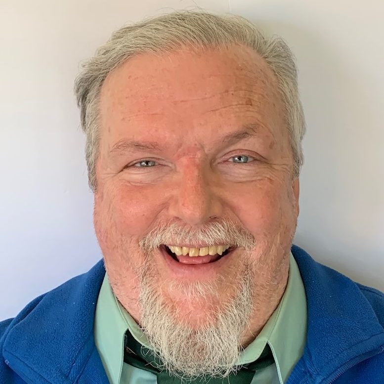 David W. Oaks: Older white male in blue vest, light green shirt and dark green tie. Short grey hair & beard, smiling.